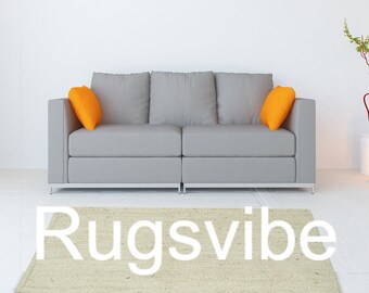 May include: A light beige rug with the text "Rugsvibe" in white lettering. The rug is in front of a gray sofa with orange throw pillows.