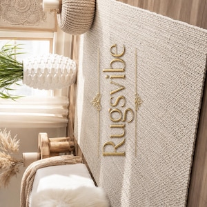May include: A rectangular area rug with a thick, woven beige texture. The word Rugsvibe is written in gold, stylized script lettering vertically across the center, flanked by two thin gold lines with decorative filigree accents on each side.