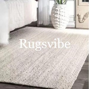 May include: A white, woven jute rug with a subtle texture. The rug is laid out on a hardwood floor and has the text "Rugsvibe" printed on it.