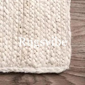 May include: White braided rug with a woven texture. The rug has a fringe border and the word "Rugsvibe" is printed on the rug.