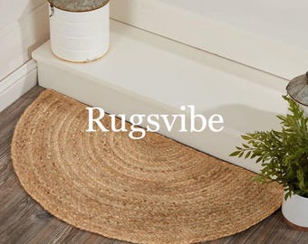 May include: A natural fiber, round, half-circle doormat made of woven jute. The doormat is placed on a wooden floor in front of a white staircase. The text "Rugsvibe" is visible in the image.