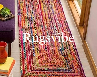 Puede incluir: Una alfombra rectangular tipo pasillero con construcción trenzada y un patrón multicolor vibrante de tiras de tela rojas, naranjas, amarillas, azules y moradas. La palabra Rugsvibe está impresa en blanco en el centro de la alfombra.
