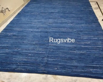May include: A large, rectangular blue rug with a textured appearance. The rug has a woven design and the text "Rugsvibe" is visible on the surface. The rug is placed on a light-coloured floor.