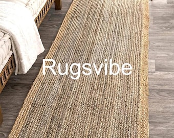 May include: A rectangular runner rug made of woven natural jute fibers, featuring a textured tan and grey center panel bordered by a thick, braided tan jute edge. The word Rugsvibe is printed in white letters across the center of the rug.