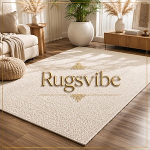 May include: A rectangular area rug with a textured, cream-colored surface. The rug features a dense, looped pile construction. Centered on the rug is the gold-colored text "Rugsvibe" flanked by two decorative gold filigree ornaments.