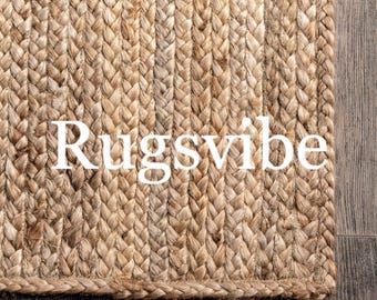 Puede incluir: Un primer plano de una alfombra de fibra natural con textura tejida. La alfombra es de color marrón claro y tiene un borde trenzado. La palabra "Rugsvibe" está impresa en blanco en la alfombra.