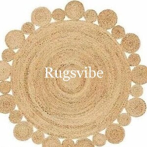 May include: A round jute rug in a natural beige colour. The rug has a circular pattern of woven jute rope around the edge. The centre of the rug is a solid beige colour. The text "Rugsvibe" is printed in the centre of the rug.
