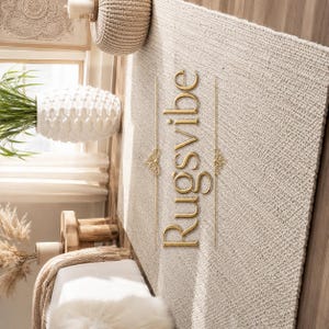 May include: A rectangular area rug with a thick, woven beige texture. The word Rugsvibe is written in gold, stylized script lettering vertically across the center, flanked by two thin gold lines with decorative filigree accents on each side.