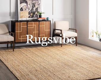 May include: A natural fiber rug with a woven texture in a light brown color. The rug is placed in a living room setting with two chairs and a wooden cabinet. The text "Rugsvibe" is visible in the center of the image.