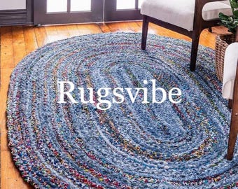 May include: A blue and white oval braided rug made from recycled denim. The rug has a unique, hand-woven design and is perfect for adding a touch of rustic charm to any room. The rug is shown in a living room setting with a white chair and a wooden floor.