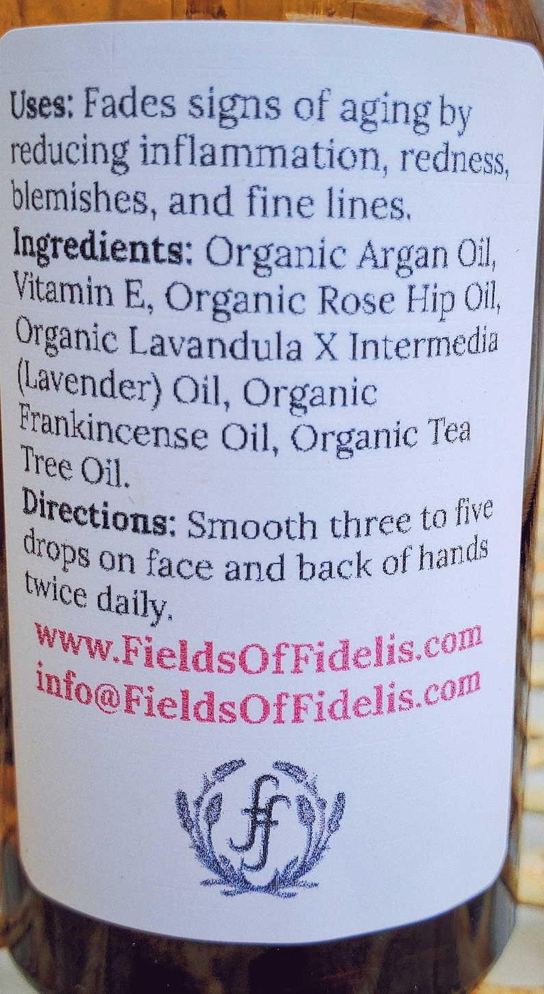 Pu&ograve; includere: Un'etichetta bianca con testo nero che descrive gli usi e gli ingredienti di un olio per il viso. L'etichetta include il nome del marchio "FieldsOfFidelis.com" e un indirizzo del sito web. L'etichetta &egrave; attaccata a una bottiglia di vetro trasparente.