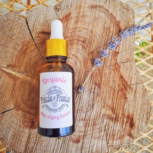 Pu&ograve; includere: Un flacone di vetro marrone con un tappo dorato e un'etichetta bianca che recita "Organic Texas Organic Fields of Fidelis Lavender Farm Anti-Aging Serum". Il flacone &egrave; appoggiato su una superficie di legno con un rametto di lavanda.
