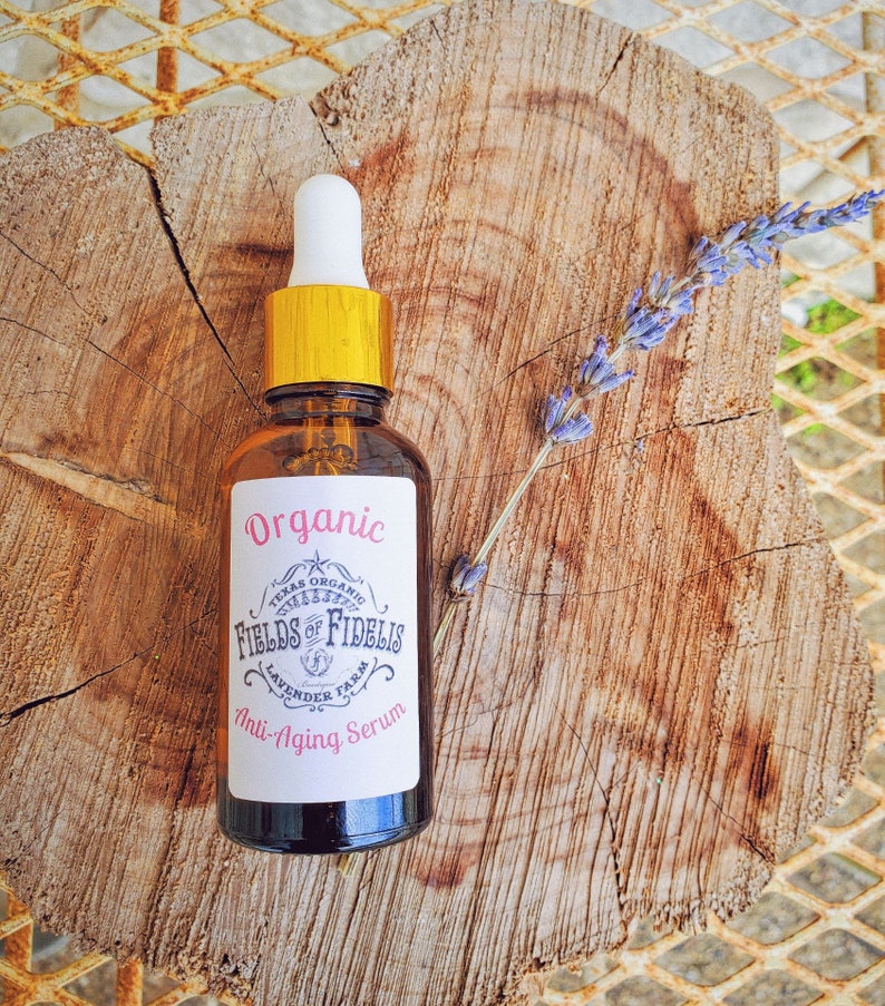 Pu&ograve; includere: Un flacone di vetro marrone con un tappo dorato e un'etichetta bianca che recita "Organic Texas Organic Fields of Fidelis Lavender Farm Anti-Aging Serum". Il flacone &egrave; appoggiato su una superficie di legno con un rametto di lavanda.