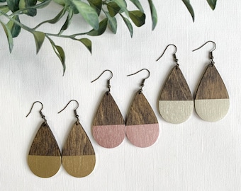 May include: Three pairs of teardrop-shaped earrings featuring a two-tone design. Each earring has a dark brown wood grain top section and a solid colored bottom section in metallic gold, dusty pink, or cream, attached to bronze-toned metal hooks.
