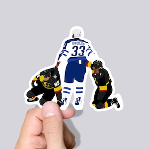 May include: A hockey sticker featuring three hockey players. The player in the centre is wearing a blue and white jersey with the name "BYFUGLIEN" and the number "33" on the back. The two players on the sides are wearing black and gold jerseys. The sticker is designed to look like a scene from a hockey game.