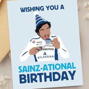 May include: A birthday card with the text "Wishing You A Sainz-ational Birthday" and an illustration of a person wearing a party hat. The card is light blue with a tan envelope.