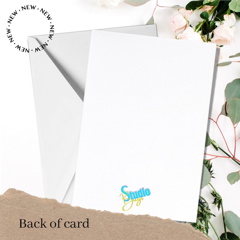May include: Blank white card with a small blue and yellow logo that says "Studio Designs" on the bottom right corner. The card is on a brown surface with white flowers and green leaves.