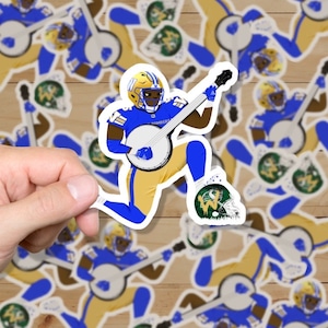 May include: A sticker featuring a cartoon football player wearing a blue and gold uniform, holding a banjo and kneeling. The player is wearing a helmet with a "W" on it. The sticker is on a wooden surface with other stickers of the same design.