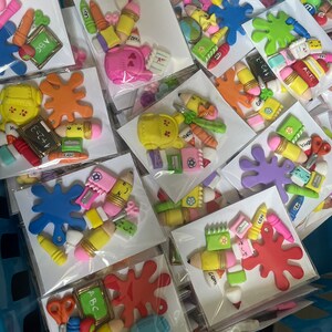 May include: A collection of colorful, handmade school-themed polymer clay charms. The charms feature various school supplies, including pencils, crayons, paintbrushes, a backpack, a ruler, and a chalkboard. The charms are packaged in individual clear plastic bags.