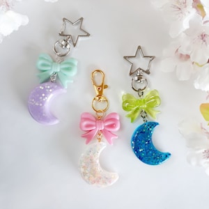 May include: Three crescent moon keychains with star-shaped charms and bows in different colors: lavender, pink, and blue.