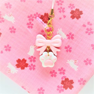 May include: A pink and white charm with a paw print and a bow. The charm is attached to a gold chain and a pink string.
