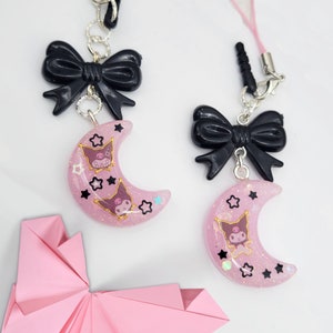 May include: Two pink crescent moon charms with black bows and a glittery finish. Each charm features a cartoon character with a pink nose and black eyes. The charms are attached to silver chains.