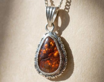 May include: A teardrop-shaped fire agate pendant set in a decorative sterling silver frame with a textured border. The gemstone displays deep orange and brown hues with internal patterns. The pendant hangs from a silver chain link necklace.