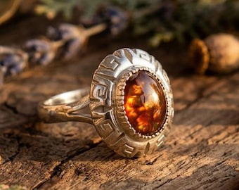 May include: A silver ring featuring an oval-shaped, translucent amber gemstone set in a bezel. The metal band is decorated with a repeating geometric Greek key pattern around the central stone, showcasing a polished finish and warm tones.