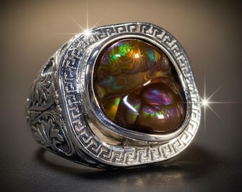 May include: A silver ring featuring a large, polished, iridescent ammolite gemstone with deep brown, green, and purple hues. The metal band is detailed with an intricate Greek key pattern around the bezel and ornate leaf carvings on the sides.