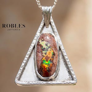 Puede incluir: Un colgante triangular de plata con una piedra central ovalada de color marrón con destellos iridiscentes verdes y naranjas. El marco de metal tiene un borde texturizado y una reasa. El texto ROBLES JOYERÍA aparece impreso en negro a la izquierda.