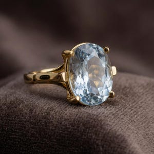 Puede incluir: Un anillo de oro con una gran gema ovalada de color azul claro. La banda y el engaste del anillo son de color oro cálido, y la gema está engastada con cuatro garras. El anillo se muestra sobre una superficie marrón.