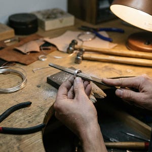 Op de afbeelding: Een zilveren ring die wordt gevijld met een metalen handvijl. De werkplek bevat een spoel zilverdraad, twee hamers met houten handvatten, een zwarte tang en stukjes leer op een houten werkbank onder het warme licht van een bureaulamp.