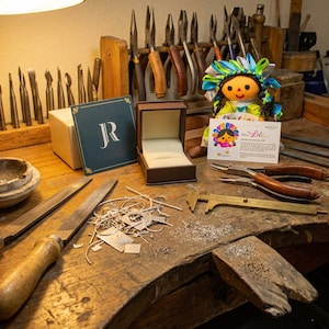 May include: A jeweler's workbench featuring a dark brown ring box, a blue card with the letters JR, a small doll, a brass caliper, metal files, and metal shavings. The doll holds a white card with the text Meet Lele and additional text.