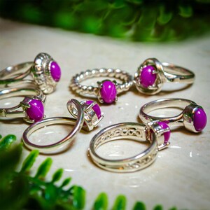 May include: Several silver rings, each set with a bright magenta oval gemstone. The rings display diverse designs, such as a twisted band and decorative settings. The magenta gemstones are the central feature, contrasted by the polished silver.