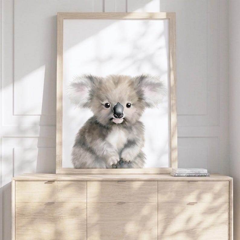May include: A framed wall art print featuring a fluffy, light brown and grey koala illustration with large, fuzzy ears, dark eyes, and a black nose, set against a plain white background, displayed in a light-colored wooden frame.