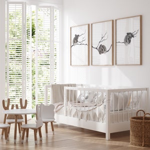 May include: A white crib with a white blanket and pillows, a white table with three white chairs, and three framed prints of koalas on a white wall.