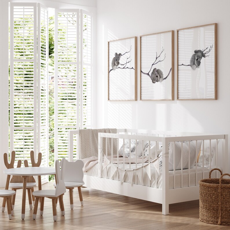 May include: A white crib with a white blanket and pillows, a white table with three white chairs, and three framed prints of koalas on a white wall.
