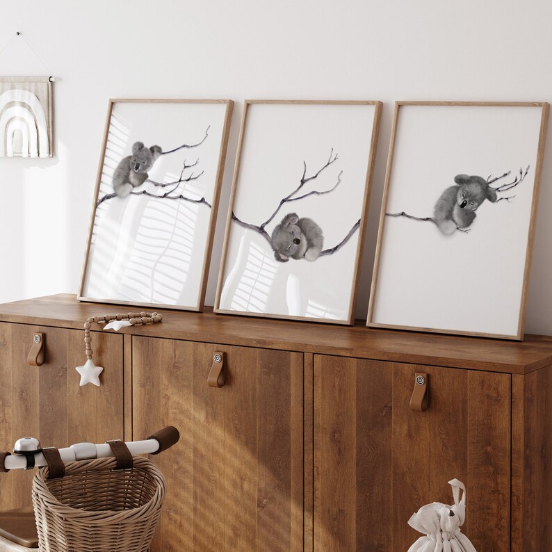 May include: Three framed prints of watercolor illustrations of koalas sleeping on branches. The prints are in a light wood frame and are hanging on a wall above a wooden cabinet.