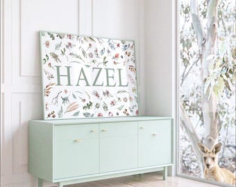 May include: A rectangular framed wall art print featuring the name HAZEL in large, sage green capital letters centered on a white background, surrounded by a botanical pattern of various leaves, twigs, and small flowers in muted earth tones.