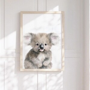 May include: A framed wall art print featuring a watercolor-style illustration of a fluffy koala with large ears, dark eyes, and a black nose. The koala is depicted in shades of gray and white, centered within a light-colored wooden frame.