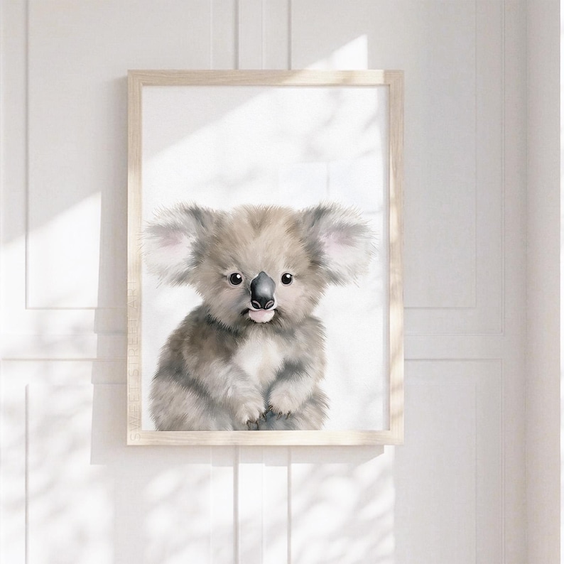 May include: A framed wall art print featuring a watercolor-style illustration of a fluffy koala with large ears, dark eyes, and a black nose. The koala is depicted in shades of gray and white, centered within a light-colored wooden frame.