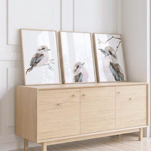 May include: Three framed wall art prints featuring watercolor illustrations of kookaburras. Each print displays a bird with brown and blue-grey plumage perched on thin branches against a white background, presented in light-colored wooden frames.
