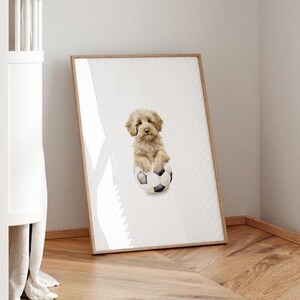 Op de afbeelding: Een ingelijste print van een lichtbruine hond die in een witte en zwarte voetbal zit. De hond kijkt naar de camera. De achtergrond is een wit en grijs gestreept patroon.