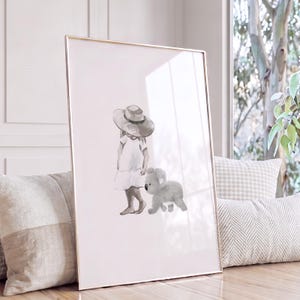 May include: A framed vertical wall art print featuring a watercolour illustration of a person wearing a wide-brimmed hat and a simple dress, walking alongside a small, fluffy grey koala. The artwork is rendered in soft, muted grey tones.