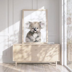May include: A framed wall art print featuring a watercolor illustration of a fluffy gray koala with large ears, a black nose, and a small pink tongue sticking out, set against a plain white background within a light-colored wooden frame.