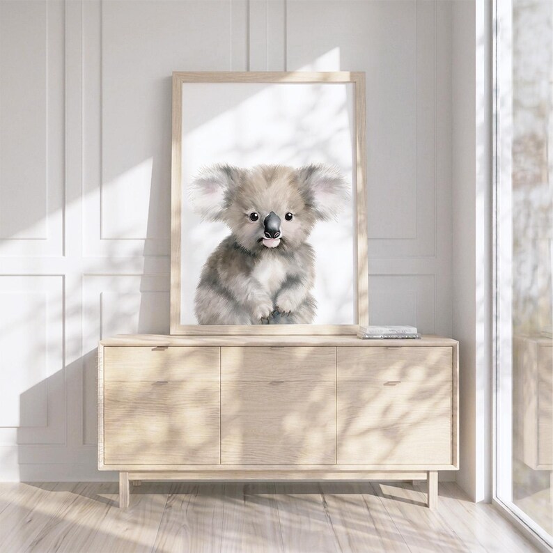 May include: A framed wall art print featuring a watercolor illustration of a fluffy gray koala with large ears, a black nose, and a small pink tongue sticking out, set against a plain white background within a light-colored wooden frame.