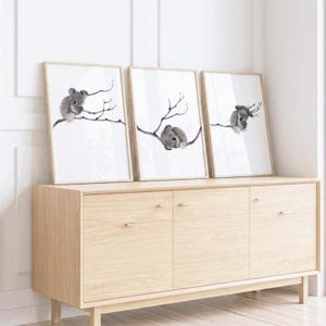 May include: Three rectangular framed wall art prints featuring black and white watercolor illustrations of koalas perched on thin, bare tree branches. Each print displays a single koala in a different pose against a plain white background.
