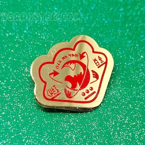 Puede incluir: Pin de metal de color dorado con un contorno rojo. El pin presenta un pez estilizado con el texto "01.2. MH. VPN" y "999" dentro del contorno.