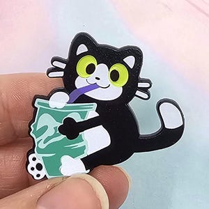 May include: A black cat-shaped charm with white paws and a white-tipped tail. The cat is drinking from a green cup with a purple straw. The cat has yellow eyes and white whiskers.