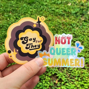 Puede incluir: Dos pegatinas con colores arcoíris y texto. La primera pegatina es marrón y amarilla con el texto "Gay for They". La segunda pegatina es iridiscente con el texto "Hot Queer Summer!"